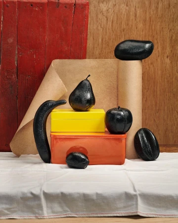 Still life of glossy black fruit and colorful boxes against a backdrop of brown paper and a red wooden panel on a white tablecloth.