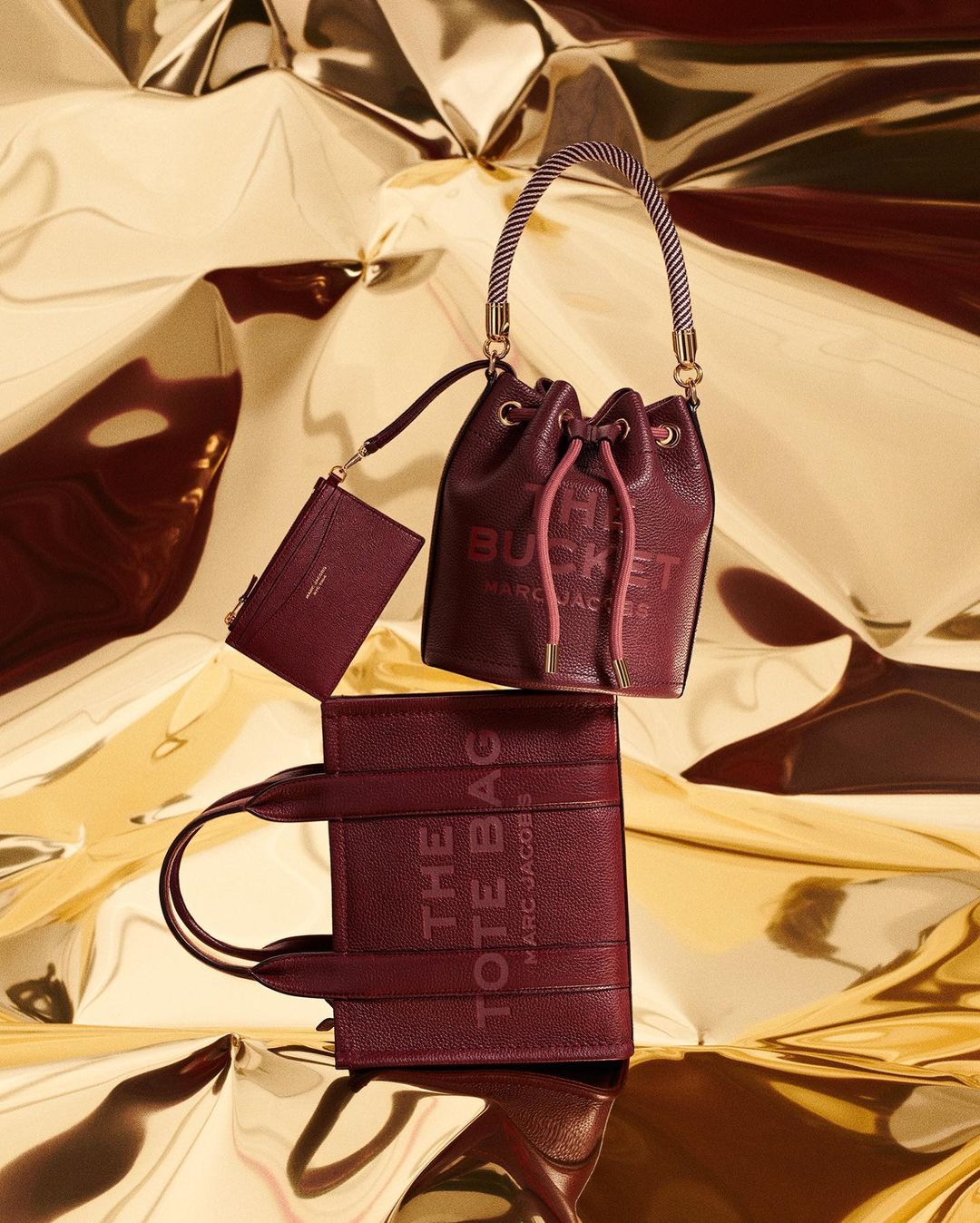 Arrangement of The Marc Jacobs burgundy leather bucket bag, tote bag, and wallet against a shiny gold background in a studio setting.