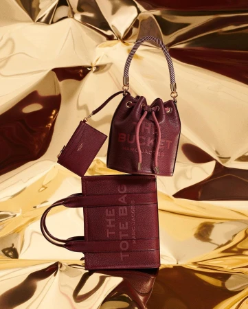 Arrangement of The Marc Jacobs burgundy leather bucket bag, tote bag, and wallet against a shiny gold background in a studio setting.