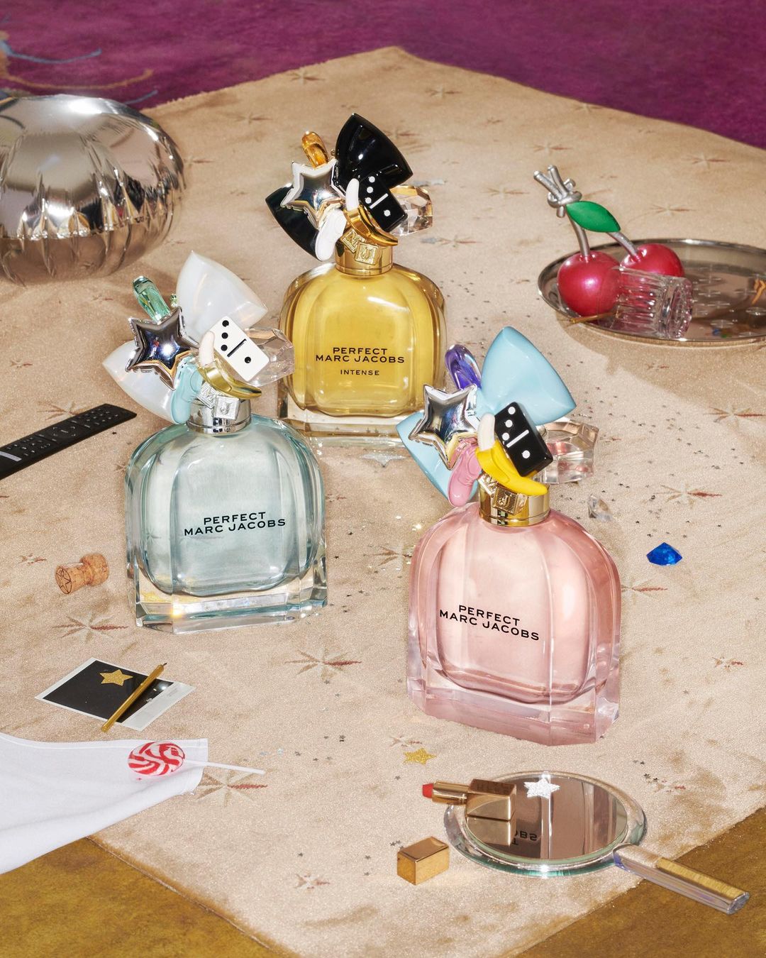 Three bottles of Perfect Marc Jacobs perfume in pink, yellow, and blue on a table with props and confetti, shot in a studio setting.