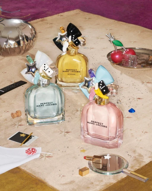 Three bottles of Perfect Marc Jacobs perfume in pink, yellow, and blue on a table with props and confetti, shot in a studio setting.