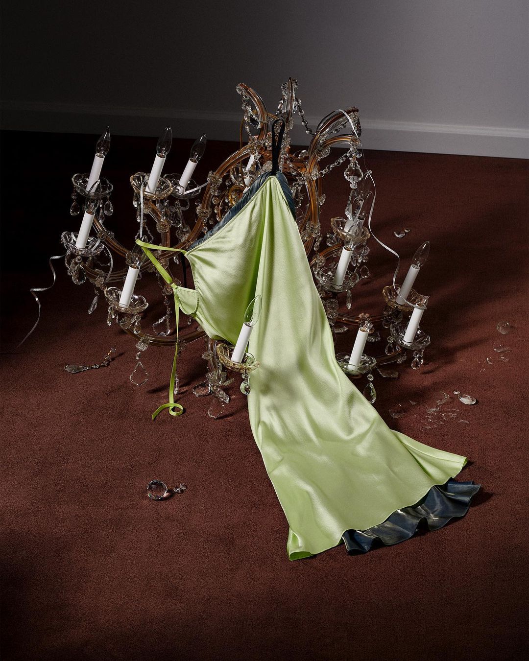 Light green satin gown draped over a vintage crystal chandelier on a brown carpet against a dark wall. High-angle, indoor shot.