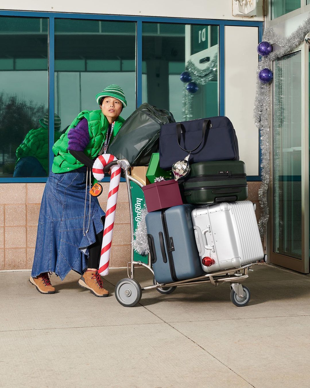 Woman with luggage cart loaded with suitcases and Christmas ornaments in front of a glass-walled airport terminal during daytime.