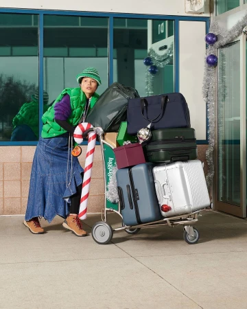 Woman with luggage cart loaded with suitcases and Christmas ornaments in front of a glass-walled airport terminal during daytime.