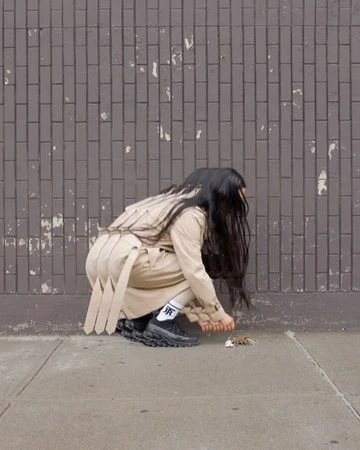 Woman in beige trench coat and sneakers crouching by a brick wall, photographed by Corey Olsen in London.