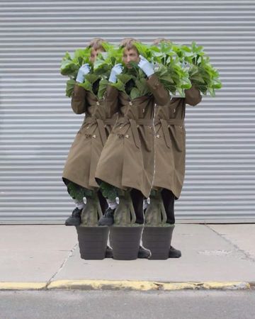 Three people wearing trench coats and plant pots stand in front of a gray corrugated metal background in an outdoor urban environment.