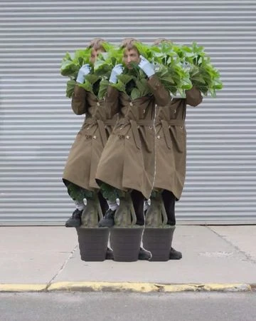 Three people wearing trench coats and plant pots stand in front of a gray corrugated metal background in an outdoor urban environment.