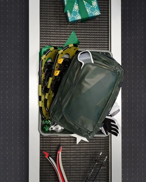 Overhead shot of green backpack, wrapped gift, snowshoes, gloves, and skis arranged in a container, set against a dark speckled backdrop.