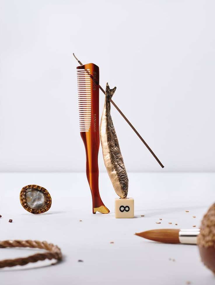 Abstract still life composition: comb with leg detail, fish, tile with '8', brush, rope circle, and small dots on a white background.