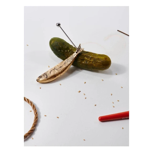 Studio shot: A golden fish figurine rests on a green pickle, accessorized with a silver cocktail pick, creating a playful, artistic composition.