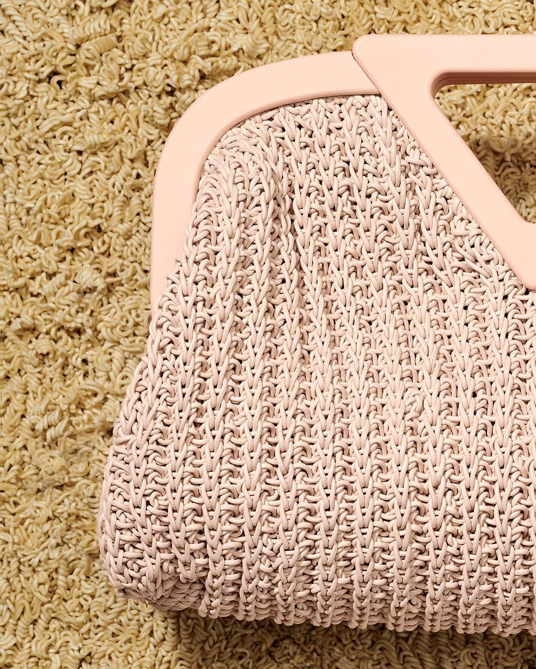 Close-up of a pastel pink woven handbag with a hard plastic handle on a textured tan background, displaying fashion and design.
