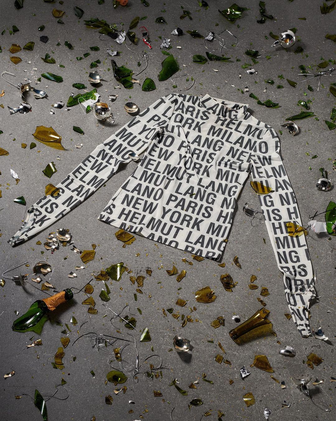 Scattered broken champagne bottle pieces and glass surround a white long sleeve shirt patterned with black text on a gray surface.