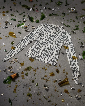 Scattered broken champagne bottle pieces and glass surround a white long sleeve shirt patterned with black text on a gray surface.