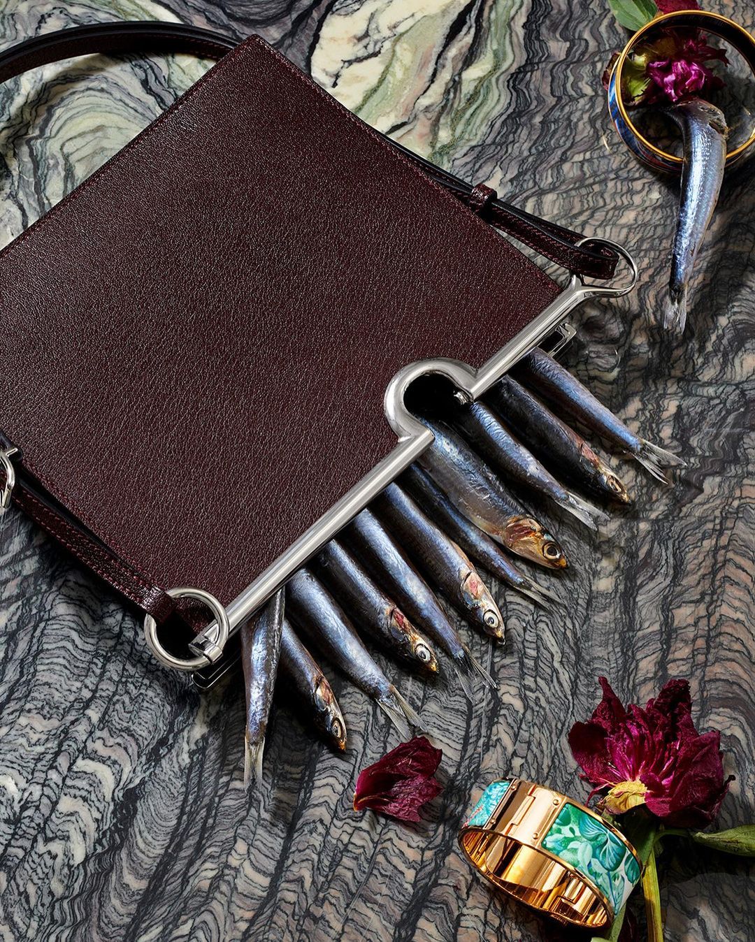 Leather purse filled with fresh fish, artfully arranged on a textured surface with floral and jewelry accents, editorial fashion.