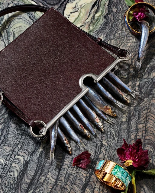 Leather purse filled with fresh fish, artfully arranged on a textured surface with floral and jewelry accents, editorial fashion.