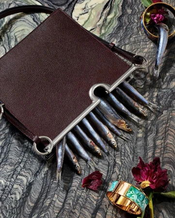 Leather purse filled with fresh fish, artfully arranged on a textured surface with floral and jewelry accents, editorial fashion.