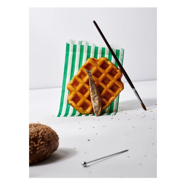 Studio shot of a waffle with a decorative sardine, a paintbrush, and a green striped bag on a white background with a chocolate pastry.