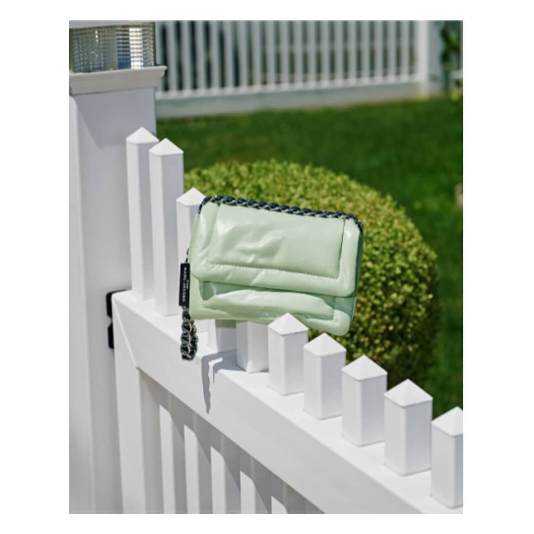 Light green puffy bag with a chain handle sits on a white picket fence in front of a green bush on a bright, sunny day outdoors.