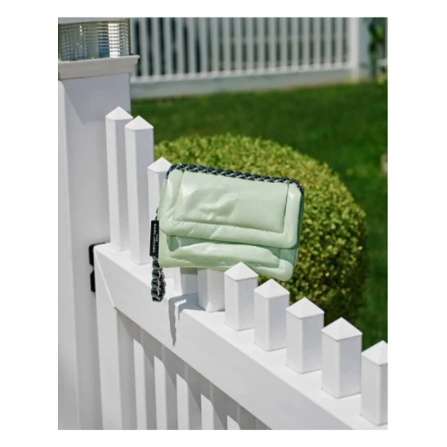 Light green puffy bag with a chain handle sits on a white picket fence in front of a green bush on a bright, sunny day outdoors.