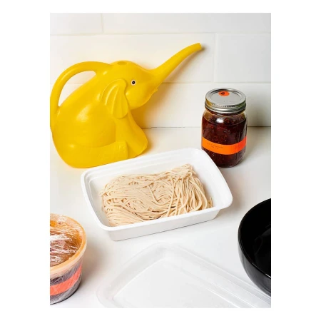 Still life on white countertop with yellow elephant watering can, noodles in a container, and jars.
