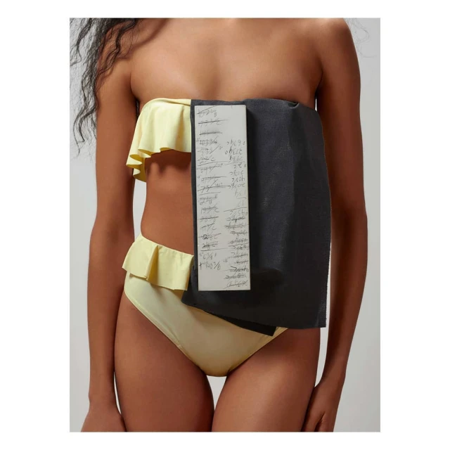 A woman wearing a yellow ruffled bikini poses with a white board with writing on it and a gray piece of fabric in a studio setting.