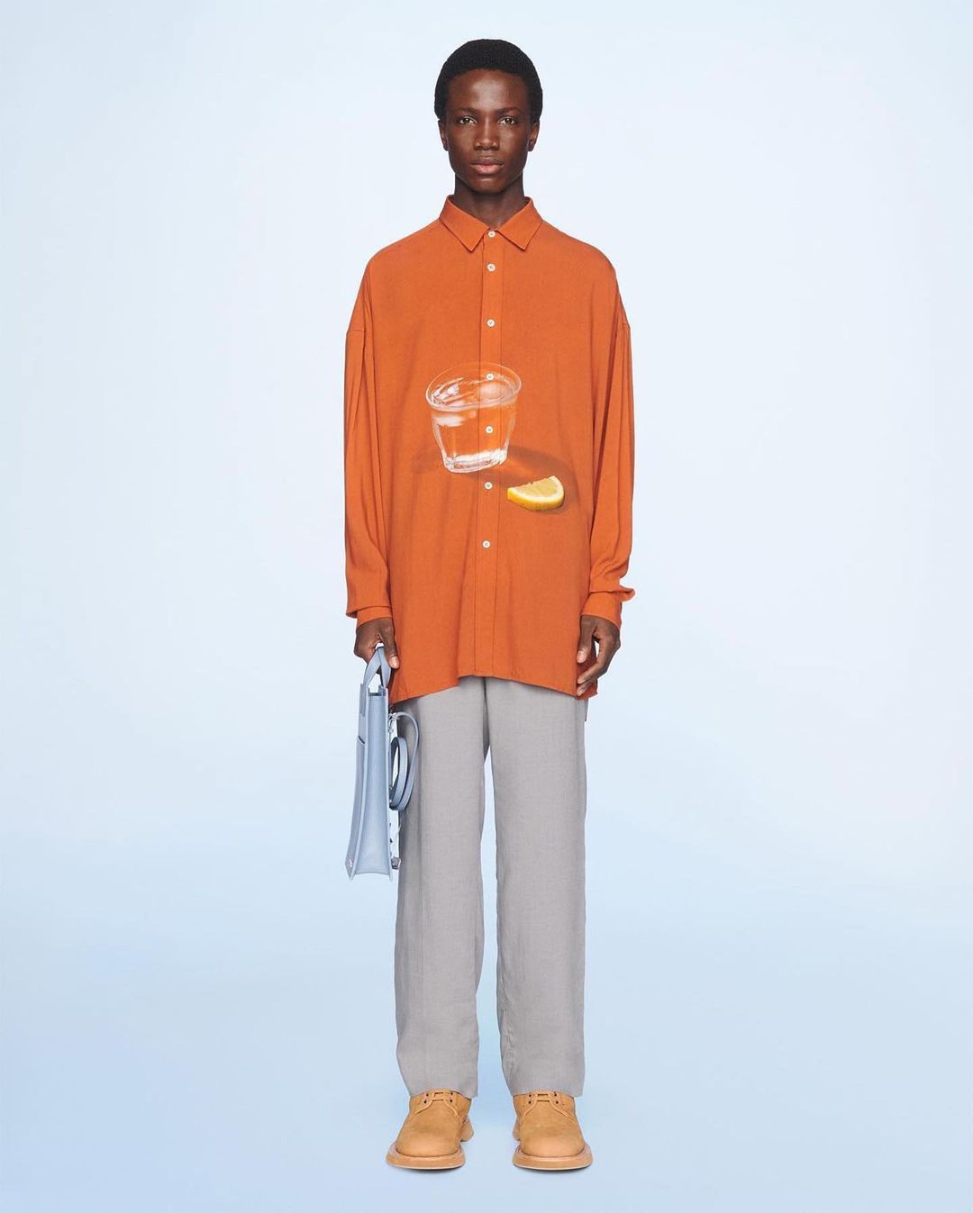 Front view of a black male model in an orange long-sleeve shirt with a glass of water and lemon, grey pants, and brown shoes against light blue.