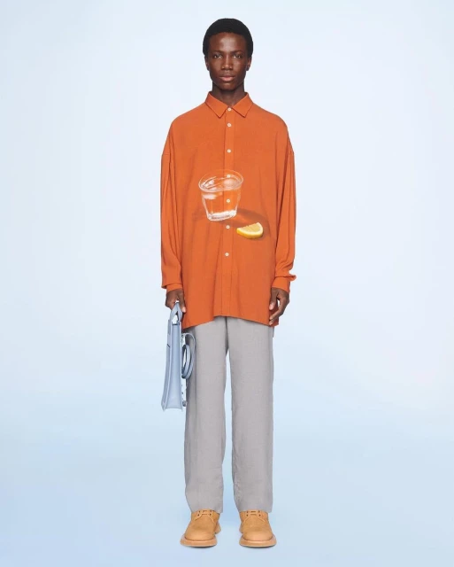 Front view of a black male model in an orange long-sleeve shirt with a glass of water and lemon, grey pants, and brown shoes against light blue.