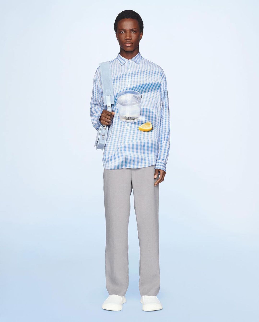 Fashion shot of an African American male model wearing a blue-and-white checkered shirt with grey pants against a light blue backdrop.