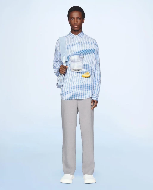 Fashion shot of an African American male model wearing a blue-and-white checkered shirt with grey pants against a light blue backdrop.