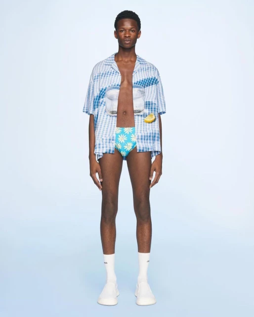 A slender man posing in an open, patterned shirt, swimsuit bottoms, and high socks set against a bright blue studio backdrop.