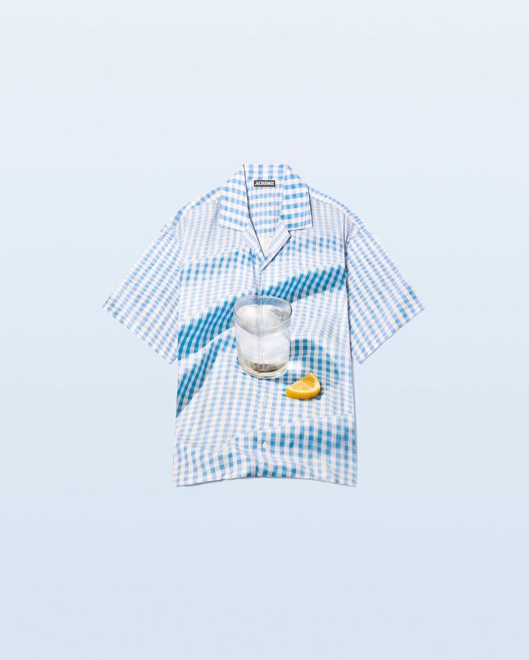 Stylized product shot featuring a blue and white gingham shirt, a clear drink in a glass, and a lemon wedge on a light blue background.