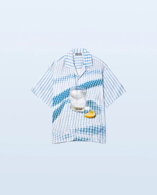 Stylized product shot featuring a blue and white gingham shirt, a clear drink in a glass, and a lemon wedge on a light blue background.