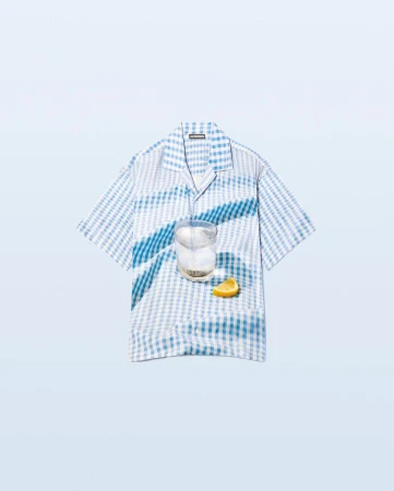 Stylized product shot featuring a blue and white gingham shirt, a clear drink in a glass, and a lemon wedge on a light blue background.