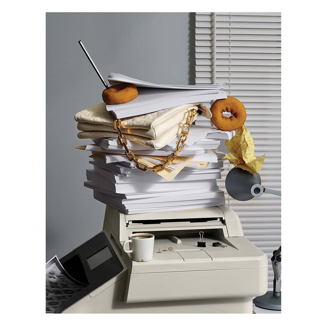 Photo of a paper pile on an office printer, topped with donuts and a handbag, against a gray wall with blinds. Coffee cup on printer.