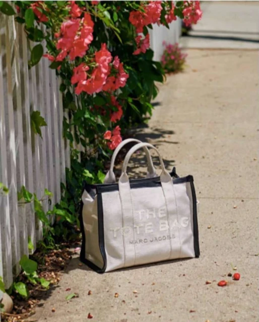 Ivory Marc Jacobs tote bag on a light gray surface near a white picket fence with pink flowers and green leaves in soft, natural lighting.