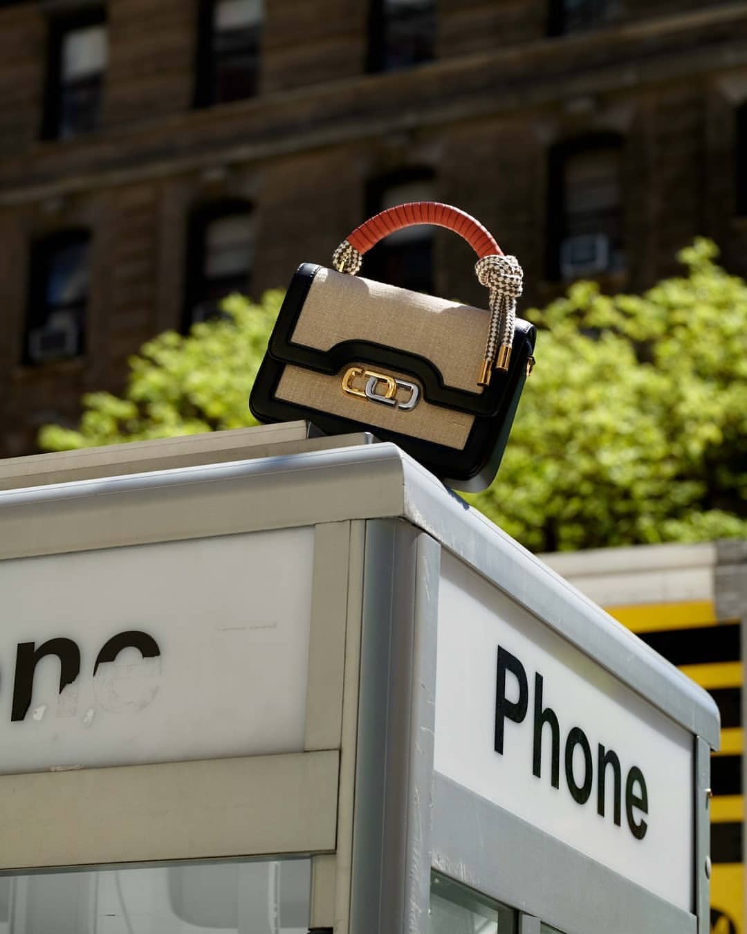 Fashionable handbag displayed on top of a gray public telephone booth, with brick building and trees in the background.