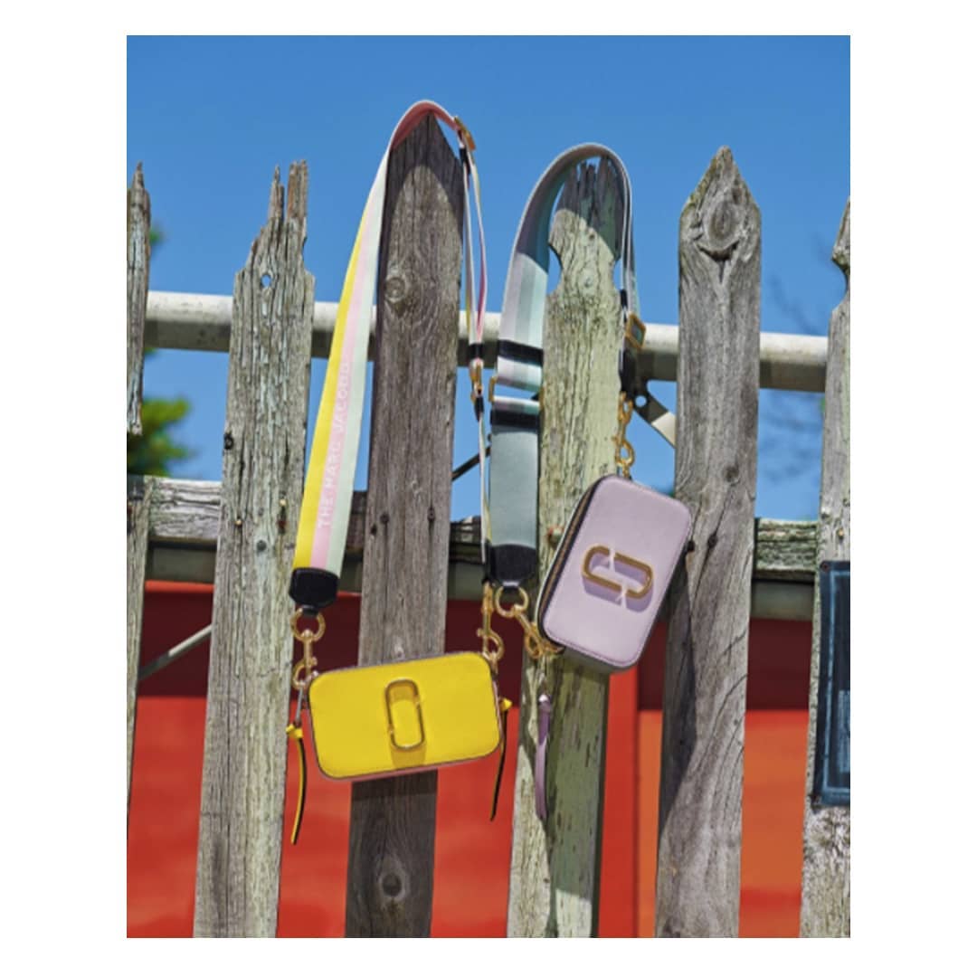 Two The Marc Jacobs snapshot bags hang from an old wooden fence against a bright blue sky. One bag is yellow, the other is lilac.