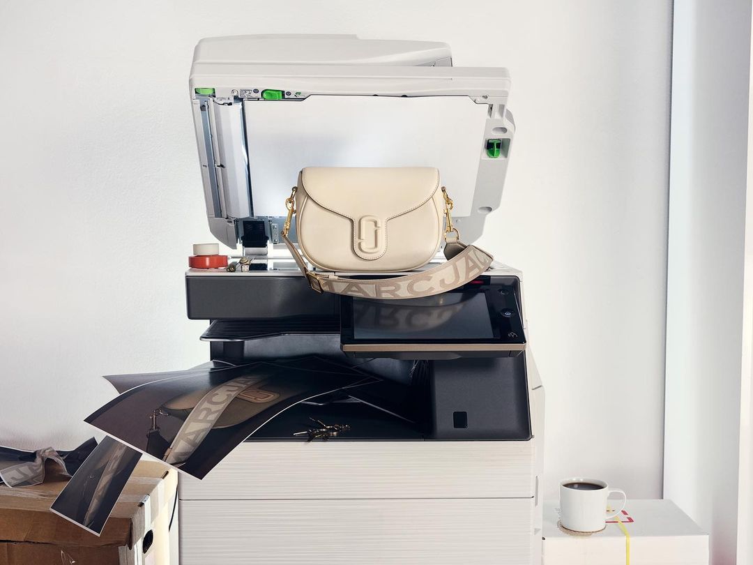 Cream-colored Marc Jacobs shoulder bag with a wide logo strap sits atop a large white office copier, with papers scattered below.