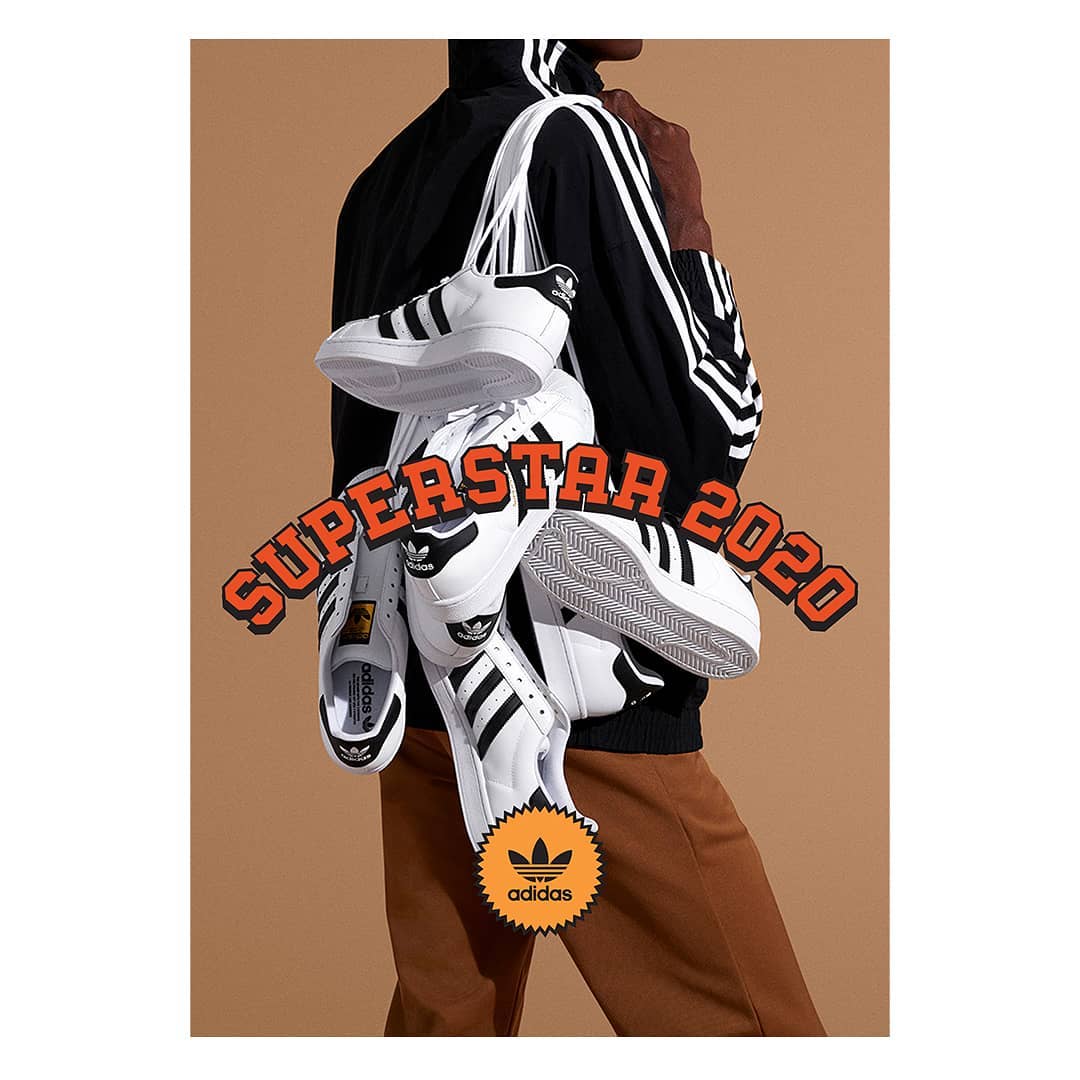 Adidas Superstar sneakers are displayed over the shoulder of a person wearing a jacket, against a tan backdrop in a product shot.