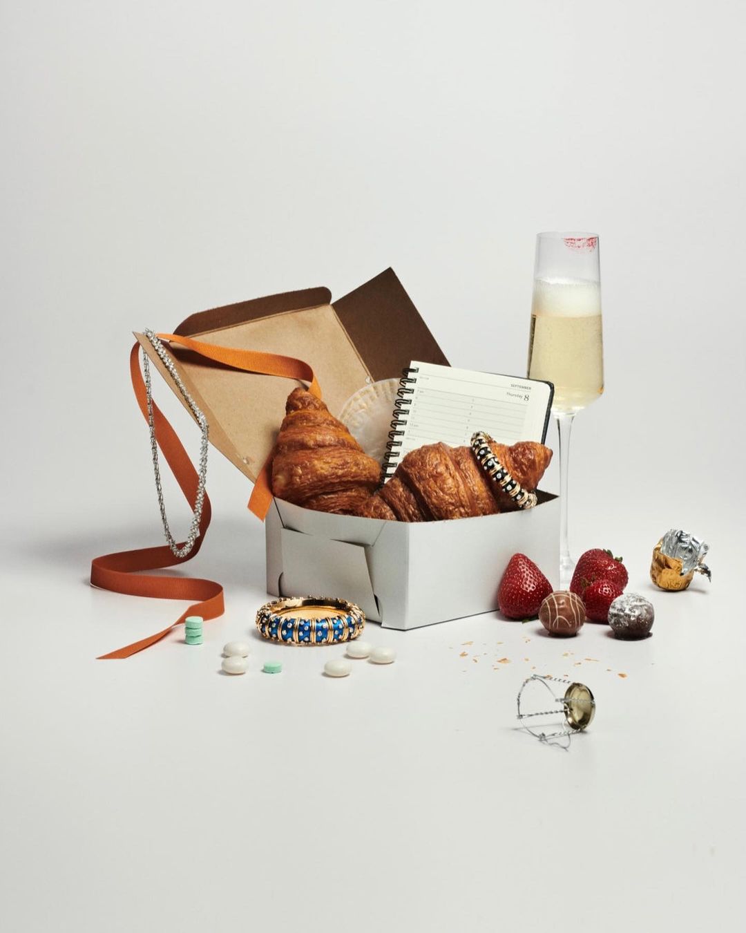 A still life arrangement features a gift box with croissants, a notebook, jewelry, champagne, and assorted sweets on a white background.