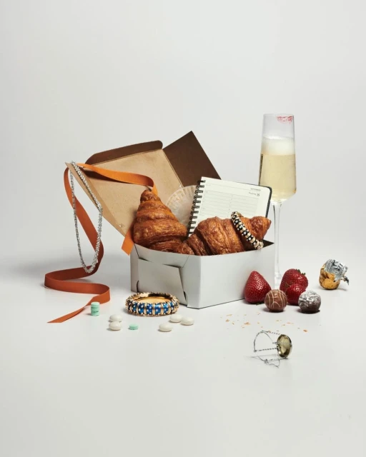 A still life arrangement features a gift box with croissants, a notebook, jewelry, champagne, and assorted sweets on a white background.