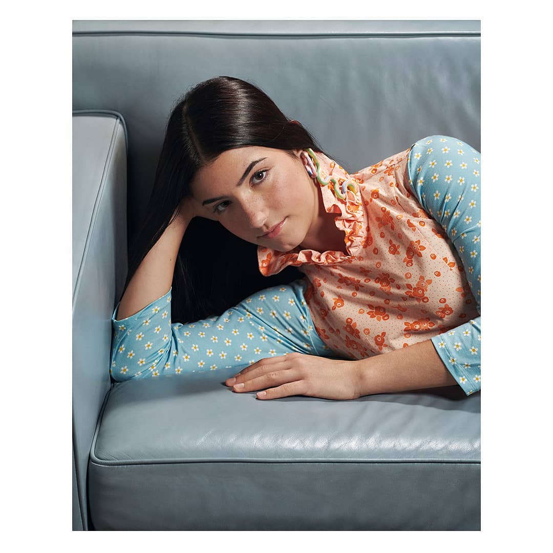 Young woman with long dark hair lies on light blue couch. She wears a long-sleeved shirt with floral patterns and a peach-colored ruffled collar.
