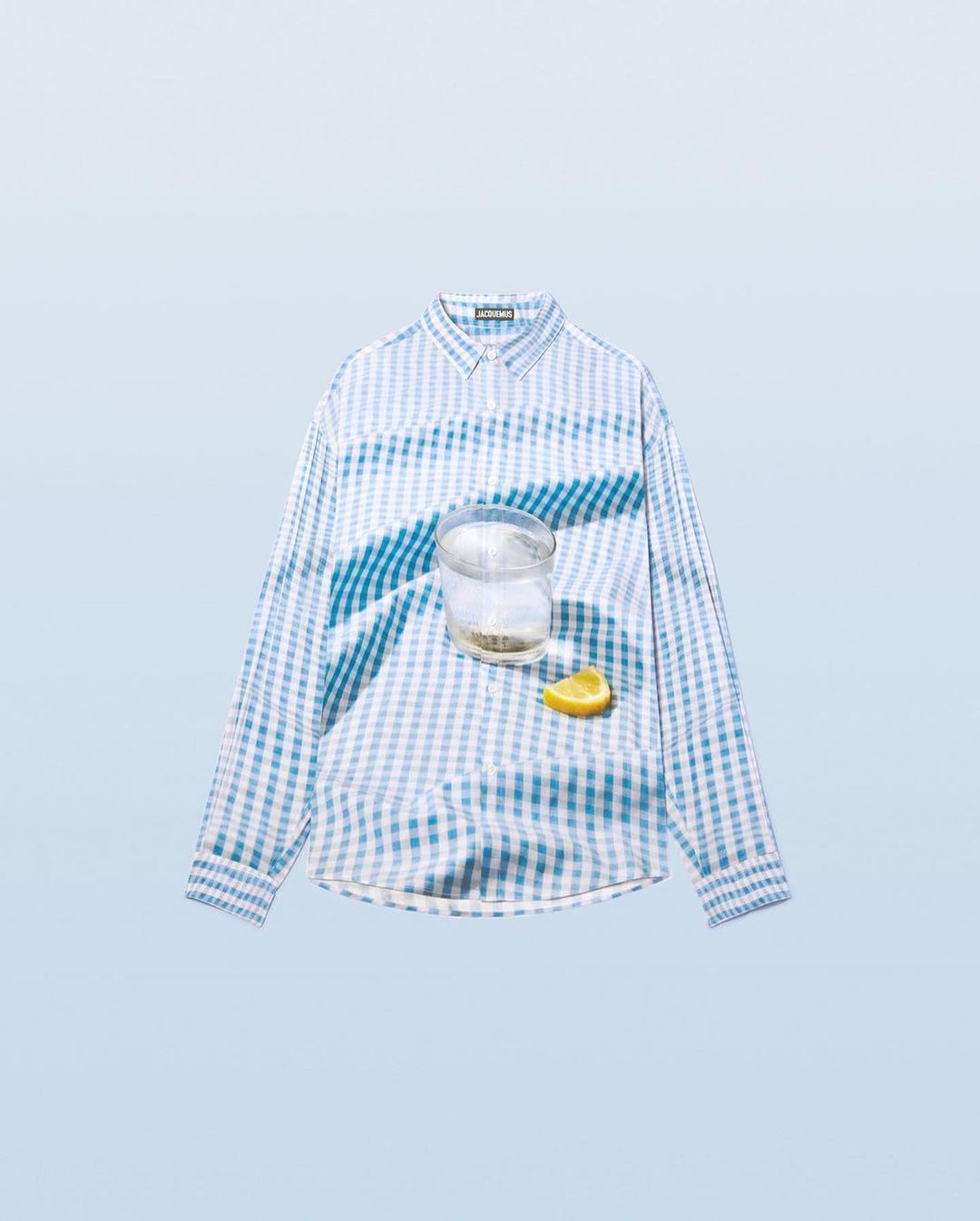 Still life flat lay: a blue and white checkered long-sleeve shirt is displayed with a glass of water and a lemon wedge, soft studio lighting.
