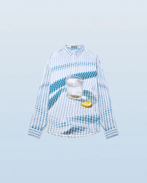 Still life flat lay: a blue and white checkered long-sleeve shirt is displayed with a glass of water and a lemon wedge, soft studio lighting.