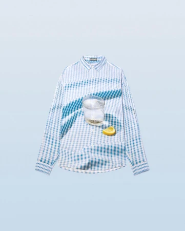Still life flat lay: a blue and white checkered long-sleeve shirt is displayed with a glass of water and a lemon wedge, soft studio lighting.