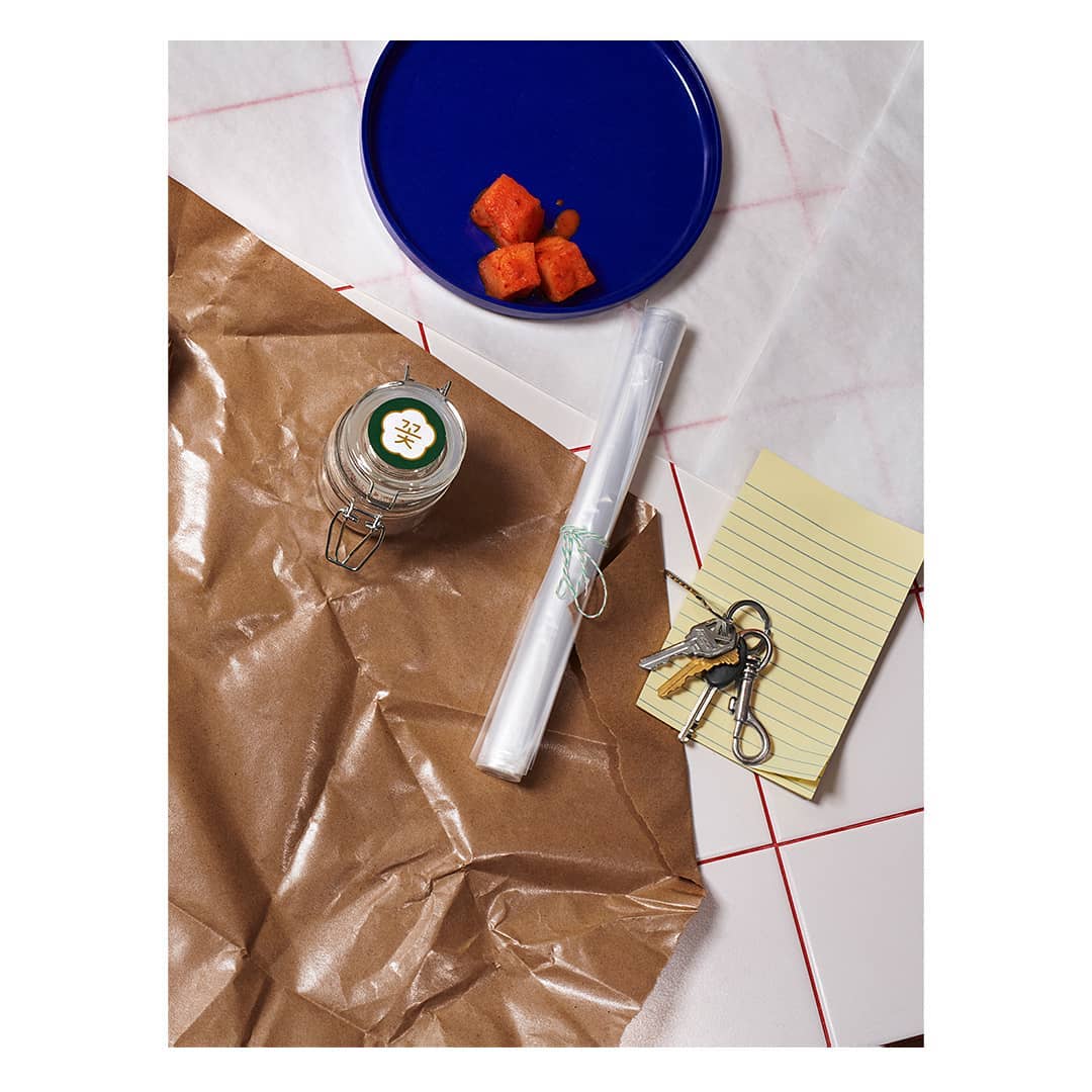 Overhead shot of a blue plate holding cubed food, a glass jar with a green and white label, cellophane wrapped object, keys, and a notepad.