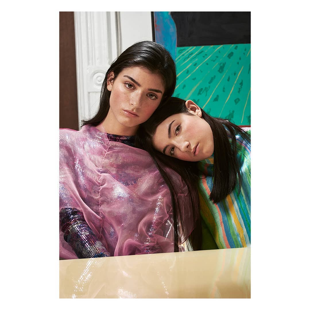 Two young women pose indoors. One wears a shiny pink top, the other a striped top. They lean against each other in a studio environment.