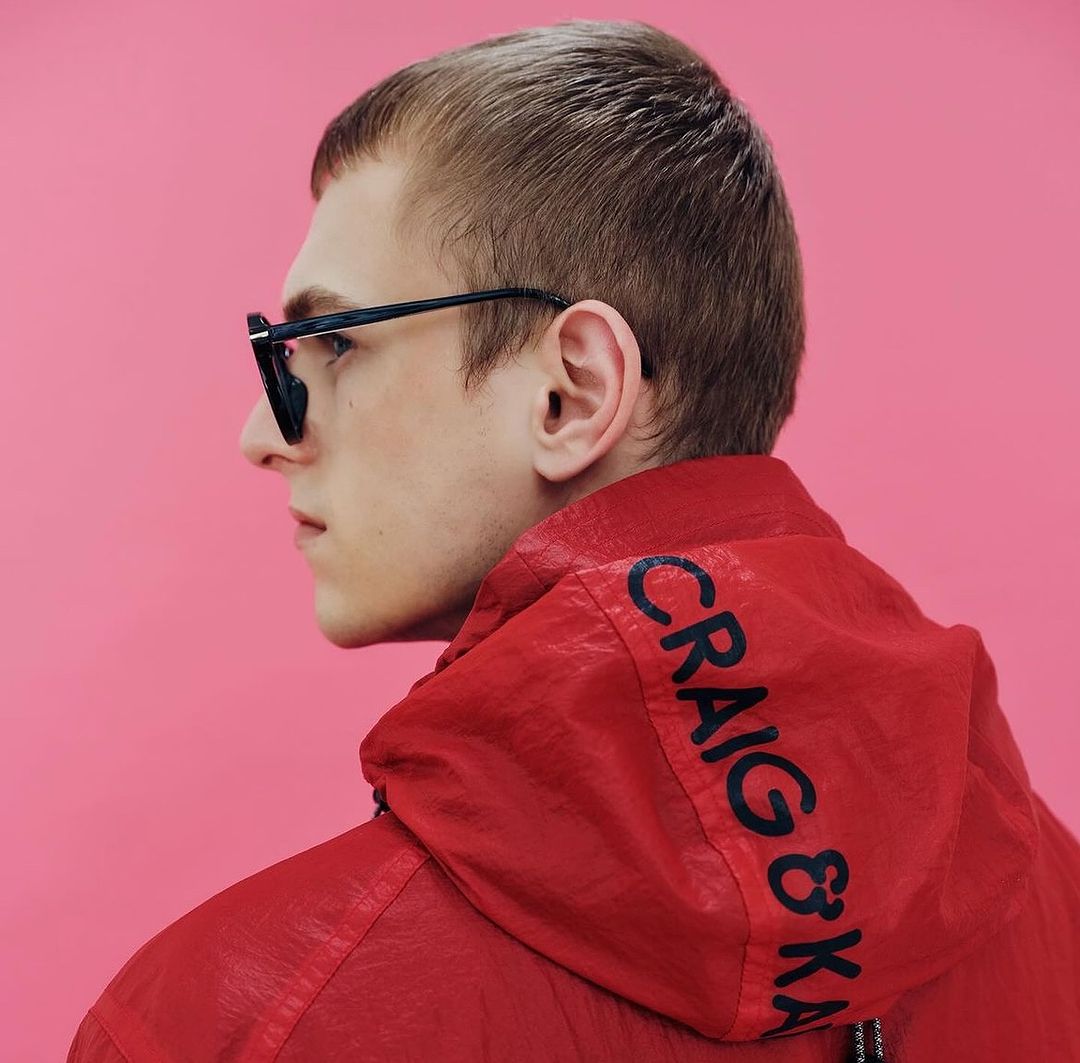 Man in profile wearing sunglasses and a red hooded jacket with the words CRAIG & KARL on the hood against a pink background.