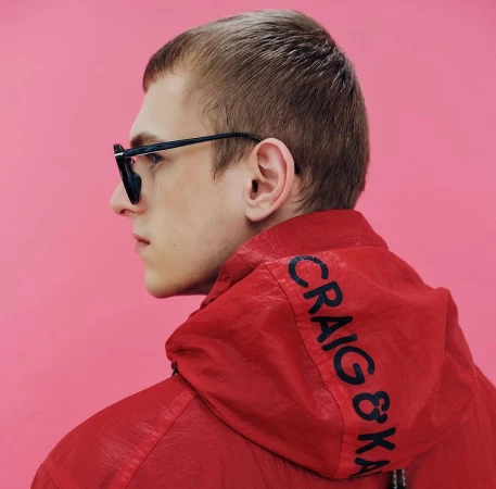 Man in profile wearing sunglasses and a red hooded jacket with the words CRAIG & KARL on the hood against a pink background.