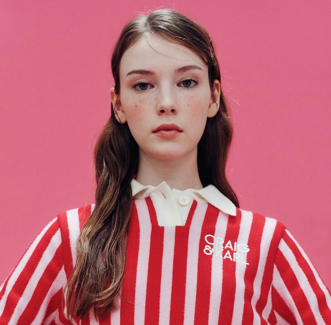 Close-up of a fair-skinned young woman with freckles, wearing a red and white striped shirt with a white collar against a pink backdrop.